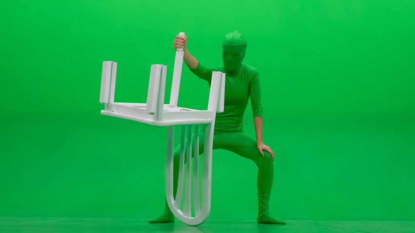 Figure in a green suit holds a white lawn chair upside down in front of green screen