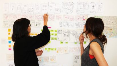 Two people standing beside a wall considering the diagrams and notes attached to it