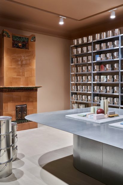 Historic tile stove on the back wall of a pale pink-hued room, with industrial shelving holding paint tins on the wall next to it