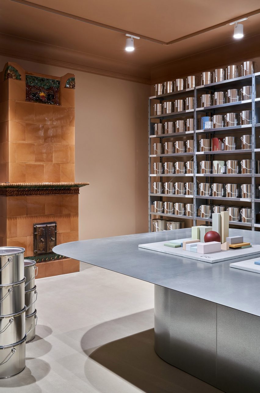 Historic tile stove on the back wall of a pale pink-hued room, with industrial shelving holding paint tins on the wall next to it