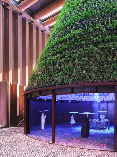 People walk with umbrellas inside a dark, rainy space underneath the green plant-covered cone