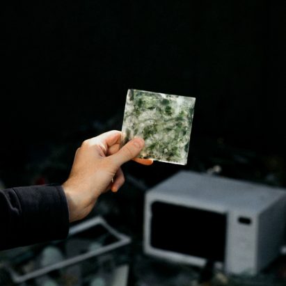 Hand holds up one Common Sands tile in front of a pile of e-waste rubbish