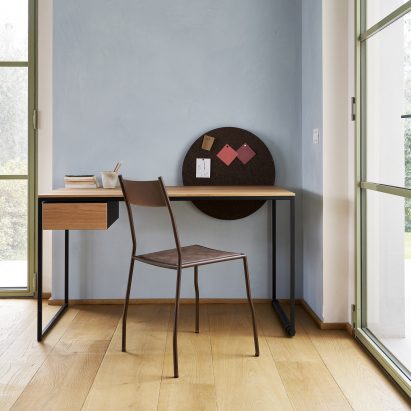 A photograph of the Macis desk in wood, set against a blue wall with a chair in front