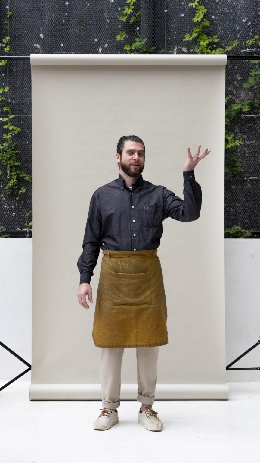 Man standing in a dark ink blue long-sleeved shirt and brown waxed apron with hand in the air