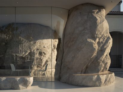 Cafe in Aranya by B.L.U.E. Architecture Studio with white chairs and tables behind glass wall that ends in rock-like column