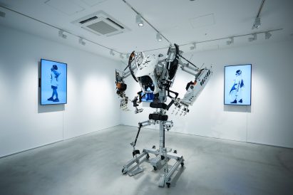 Large exoskeleton-like suit stands in the middle of a white gallery space surrounded by digital artwork on the walls in the Sai Gallery in Tokyo