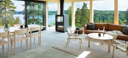 Dining and living room furnished with natural wood tables and chairs with CLT wall structure and large windows overlooking a forest and lake landscape