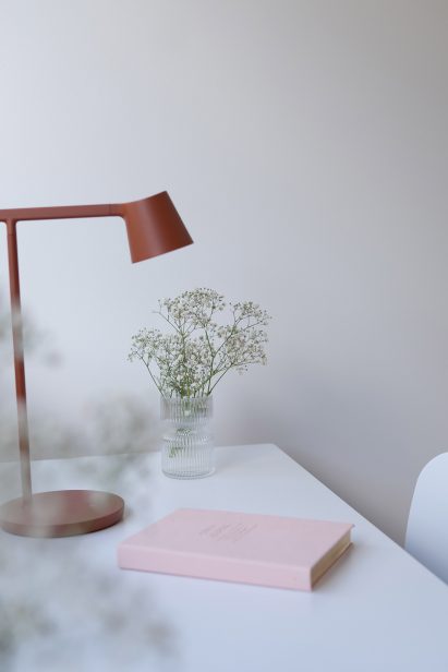 A wall painted in a cool toned mauve Teknos paint with a white desk, red desk lamp, pink diary and a jar of flowers