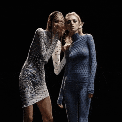 Two hologram models whispering to each other, one with pointed ears the other with slicked down hair
