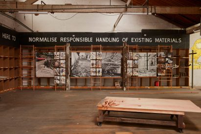 Corner of Revival Projects' construction waste hub with shelving covered in photograph display and painted sign reading 'here to normalise responsible handling of existing materials'