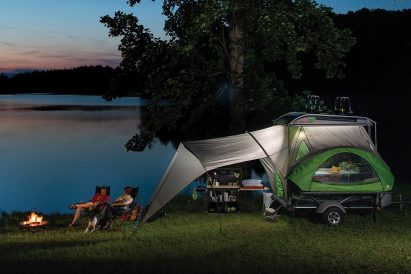Two people sit by a fire beside a pop-out tent trailer next to a lake