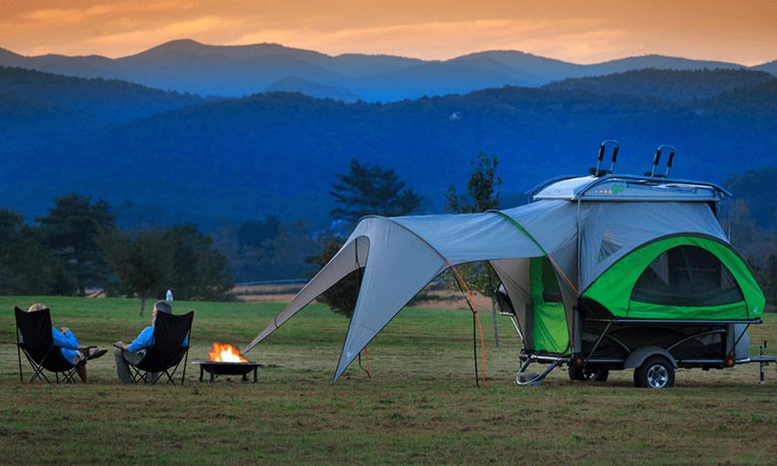Two people sit by a fire beside a pop-out tent trailer in the mountains