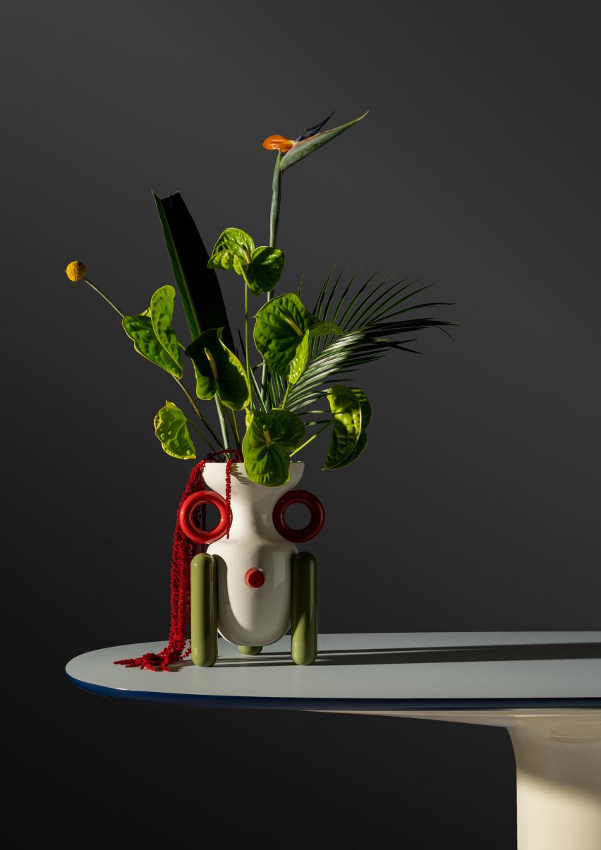 White Explorer vase with green leg stands and red handles in a showroom space with large green leafs and flowers