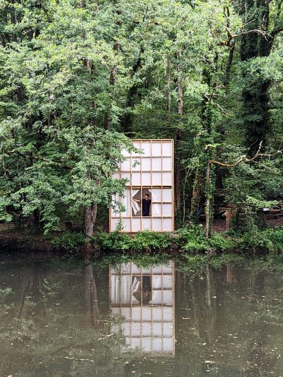 A man opening curtains inside a cabin on the edge of a lake