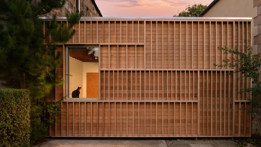Photograph showing a timber-clad structure on the side of a house at dusk/dawn with a cat in the window