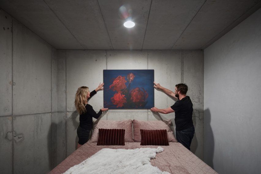 Photograph showing concrete bedroom with two people hanging art above bed