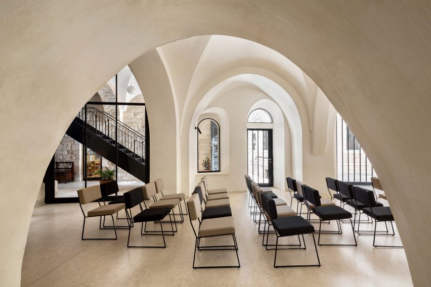 View through white arches of a room set up with rows of black-framed chairs with black window frames and staircase in the background