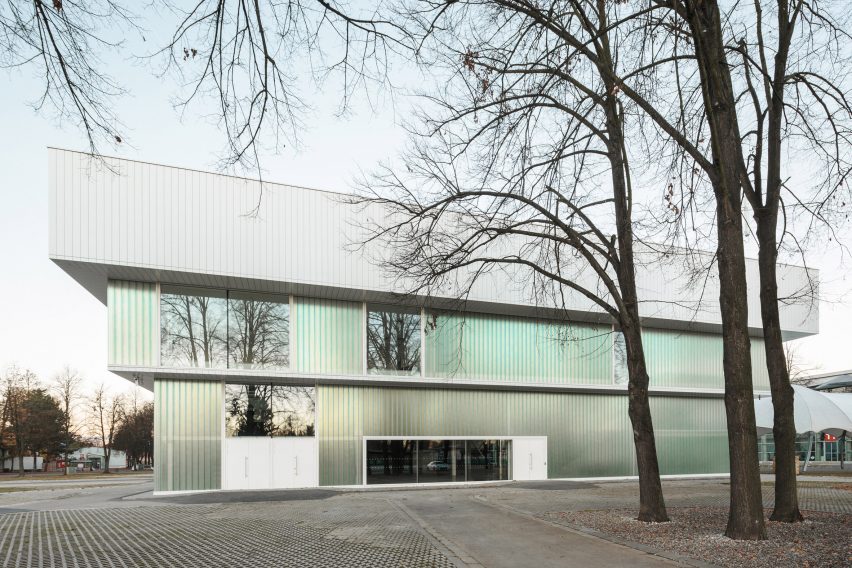 Photograph of low-slung white building with glazing with tree in foreground