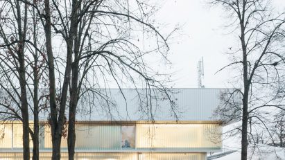 Photograph of low-slung white building with glazing with tree in foreground