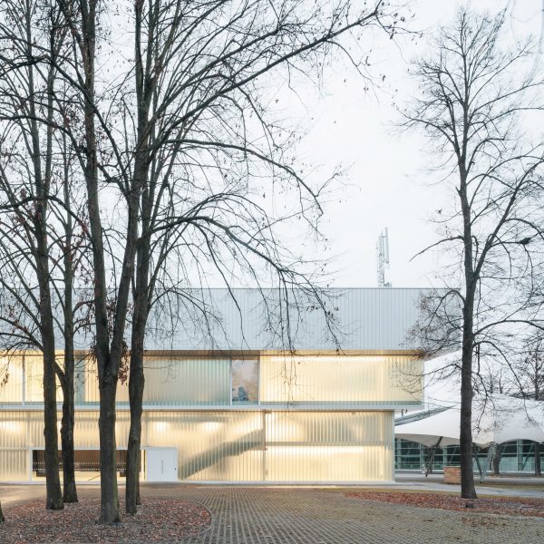 Photograph of low-slung white building with glazing with tree in foreground