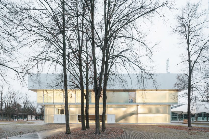 Photograph of low-slung white building with glazing with tree in foreground