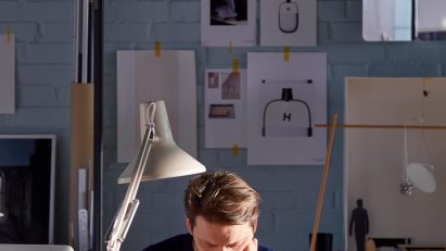 Daniel Rybakken sits at a desk in his studio, with drawings pinned to the wall behind him