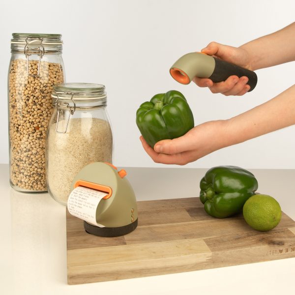 A worktop with glass jars of grains, a wooden chopping board and a person holding a device up to a green bell pepper