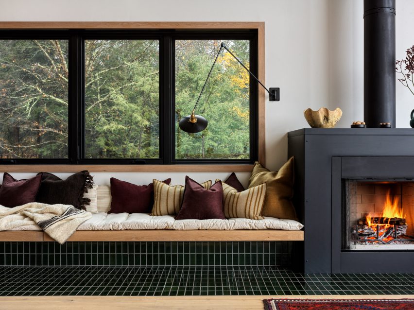 Built-in seating covered in black tiles under a window, with cream blankets and yellow and purple cushions next to a fireplace