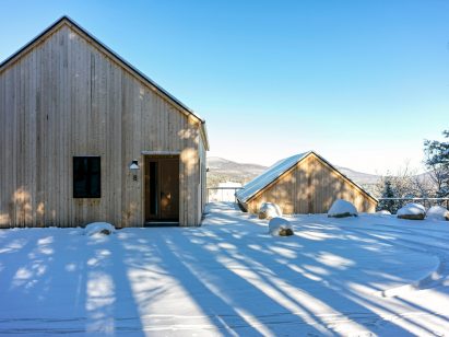 Two timber-clad pitched-roof structures on a snowy hill with trees