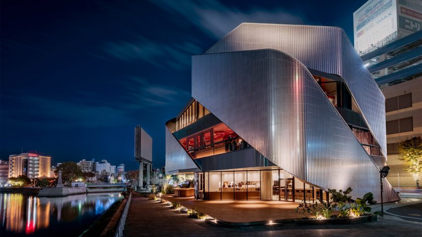 A theatre building on a city street corner with sweeping steel exterior panels lit by spotlights on the ground