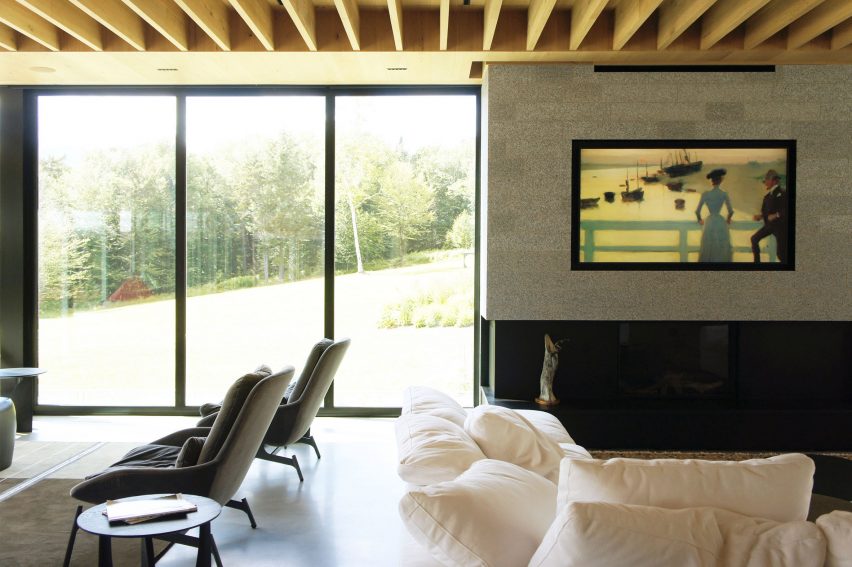 A living room with a white sofa, lounge chairs and floor-to-ceiling windows overlooking a forest