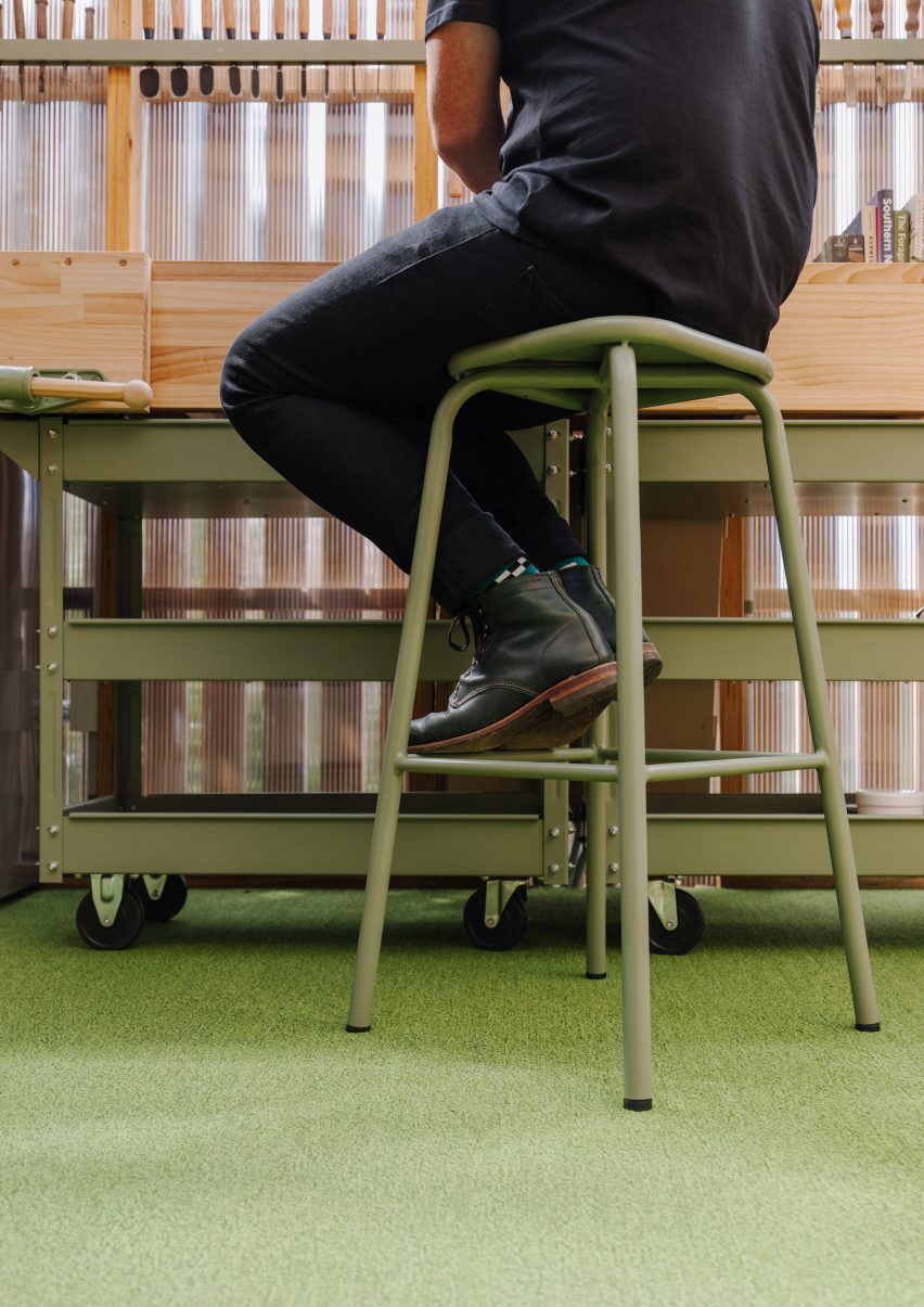 A person sitting on a green metal stool at a timber workbench, with green storage on wheels and green floor