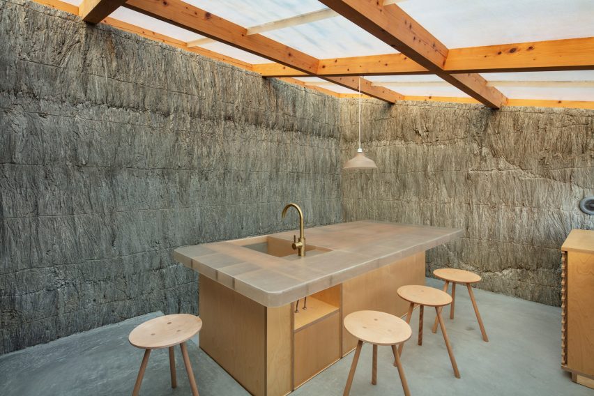 A dark kitchen space with textured stone walls, timber frame roof and large central island with a sink lit