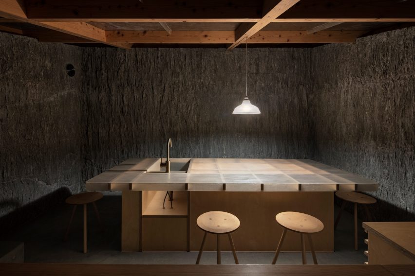 A dark kitchen space with textured stone walls, timber frame roof and large central island with a sink lit by a pendant light