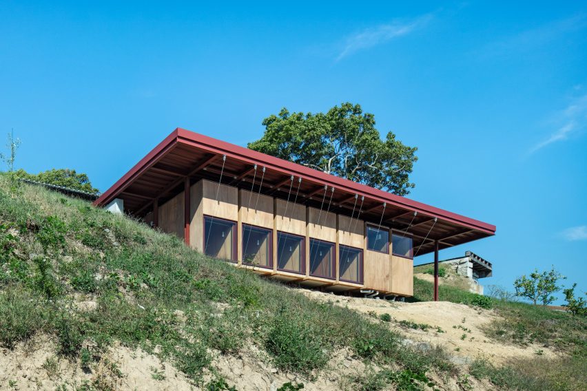 A guesthouse with large windows, wood walls and an overhanging red steel roof on a hillside