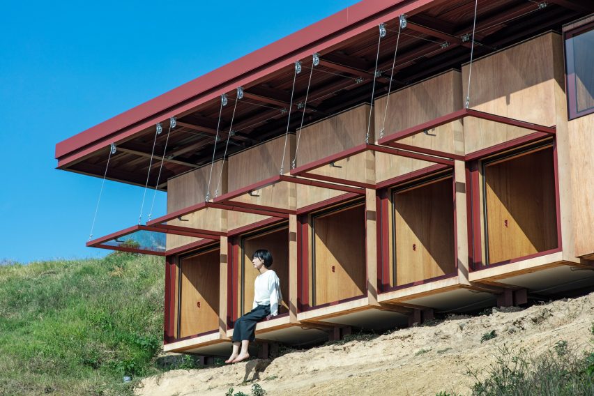A building with wood walls, red steel roof and a person sitting an opening of a series of large top-hinged windows on a hillside