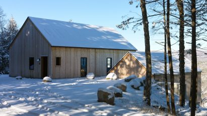 Two timber-clad pitched-roof structures on a snowy hill with trees