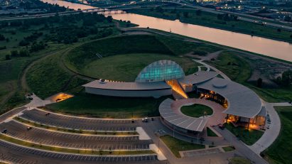 Aerial view of a curved building sunken into an earthen mound in a grassy landscape near a river