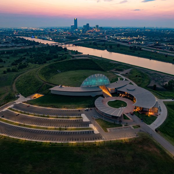 Aerial view of a curved building sunken into an earthen mound in a grassy landscape near a river