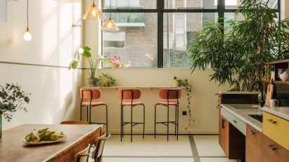 Communal kitchen and dining area with white walls and floors, large windows in dark metal frames, wood kitchen units, wood dining table and pink bar stools by The Mint List