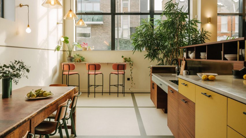 Communal kitchen and dining area with white walls and floors, large windows in dark metal frames, wood kitchen units, wood dining table and pink bar stools by The Mint List