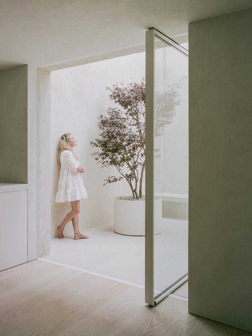 An interior space with wood floors and an pivoting glass door opening onto a white outdoor courtyard with a potted tree