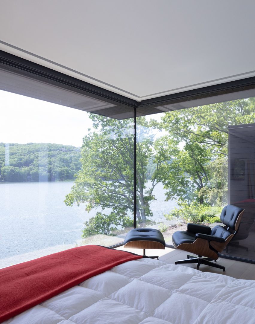 Bed, lounge chair and foot stool in a bedroom with a large corner window overlooking a lake
