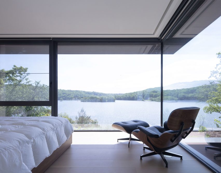 Lounge chair, foot stool and bed in a bedroom with a large corner window overlooking a lake