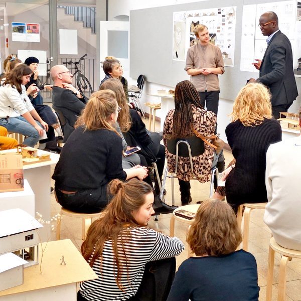 Students presenting architecture work to other students sat on wooden stools
