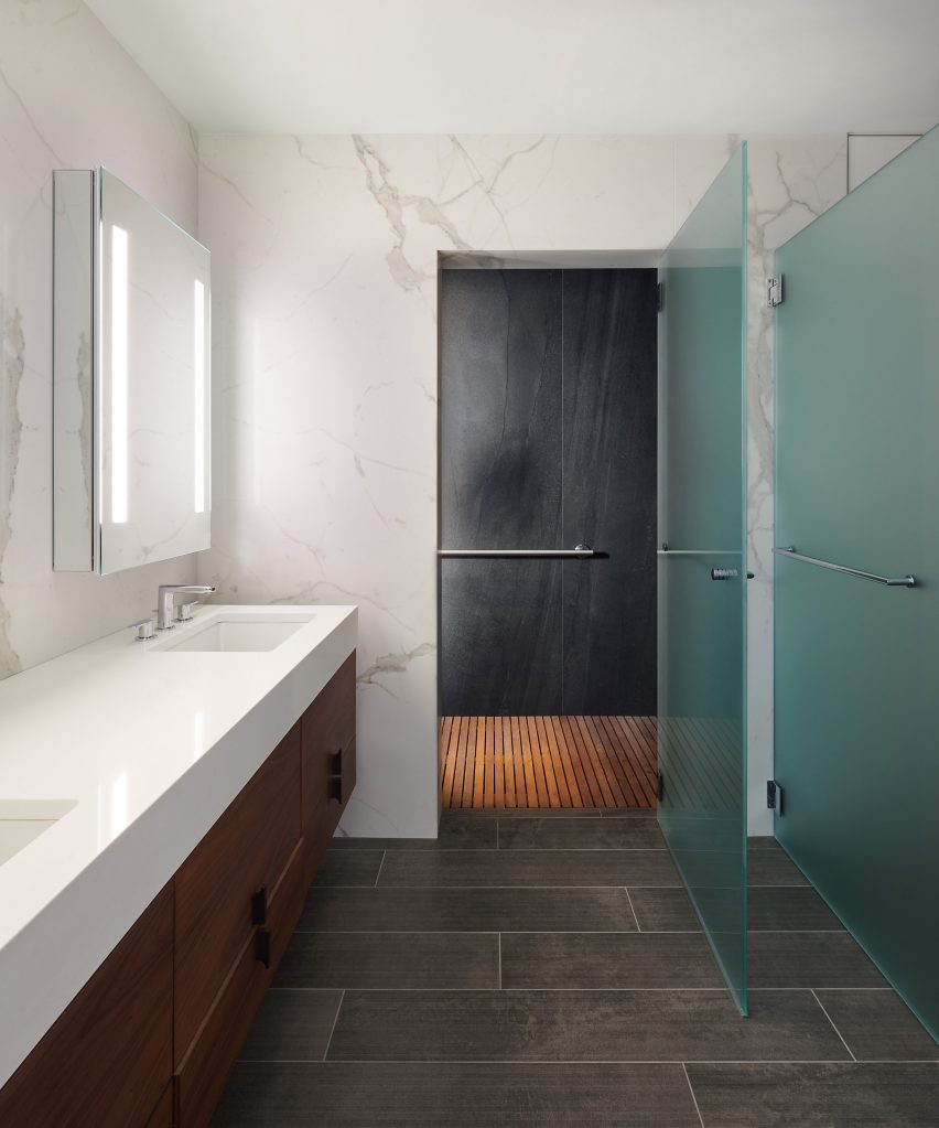 A bathroom with dark tiled flooring, white marble walls and white basins with wood drawers