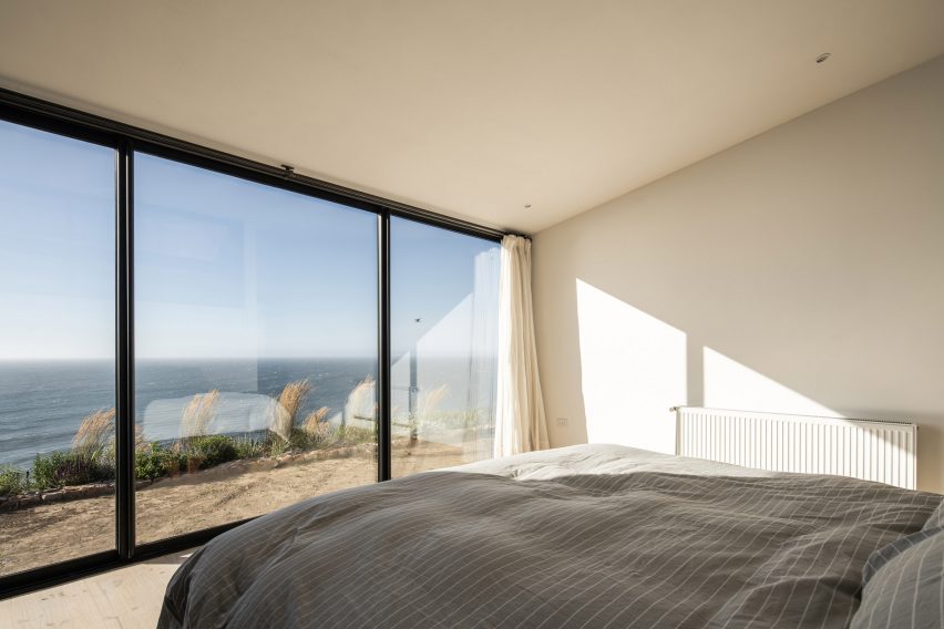 Bedroom with white walls, a bed with grey bedding and floor-to-ceiling windows overlooking the sea
