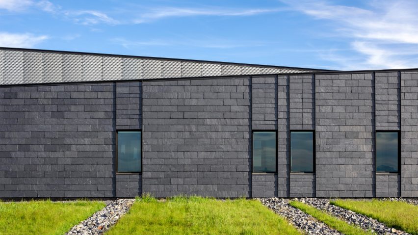 Facade of a slate-clad building with a sloping roof and a grass lawn in front