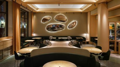 A restaurant interior with wood columns and beams, wood tables, green upholstered seating and oval metallic wall lights