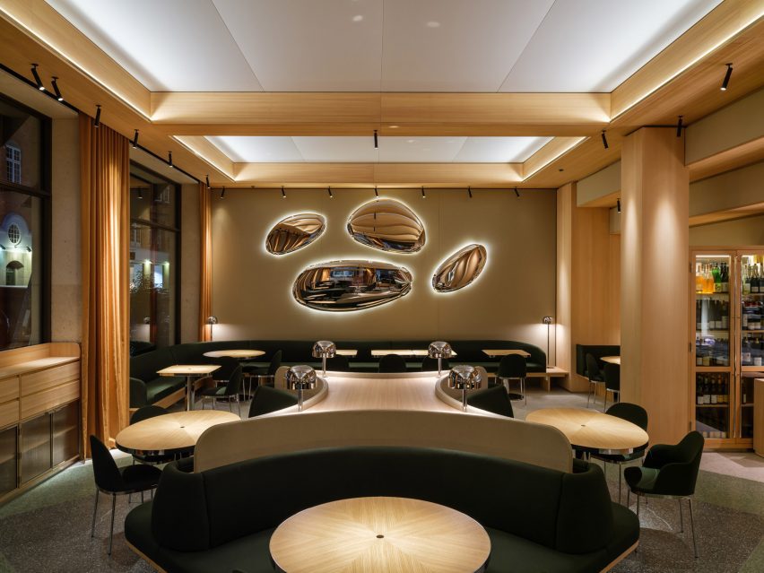 A restaurant interior with wood columns and beams, wood tables, green upholstered seating and oval metallic wall lights